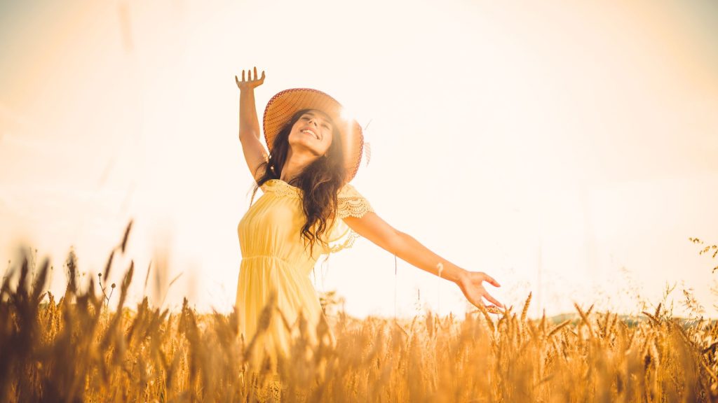 woman in straw hat and yellow sundress waving arms in a field of wheat at sunrise