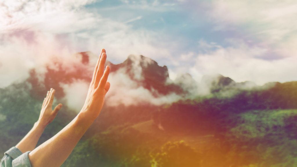 delight yourself in the Lord arms raised in worship in the foreground with mountains and clouds in the background