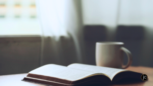 An open Bible and a steaming mug on a table in front of an open window in the morning light