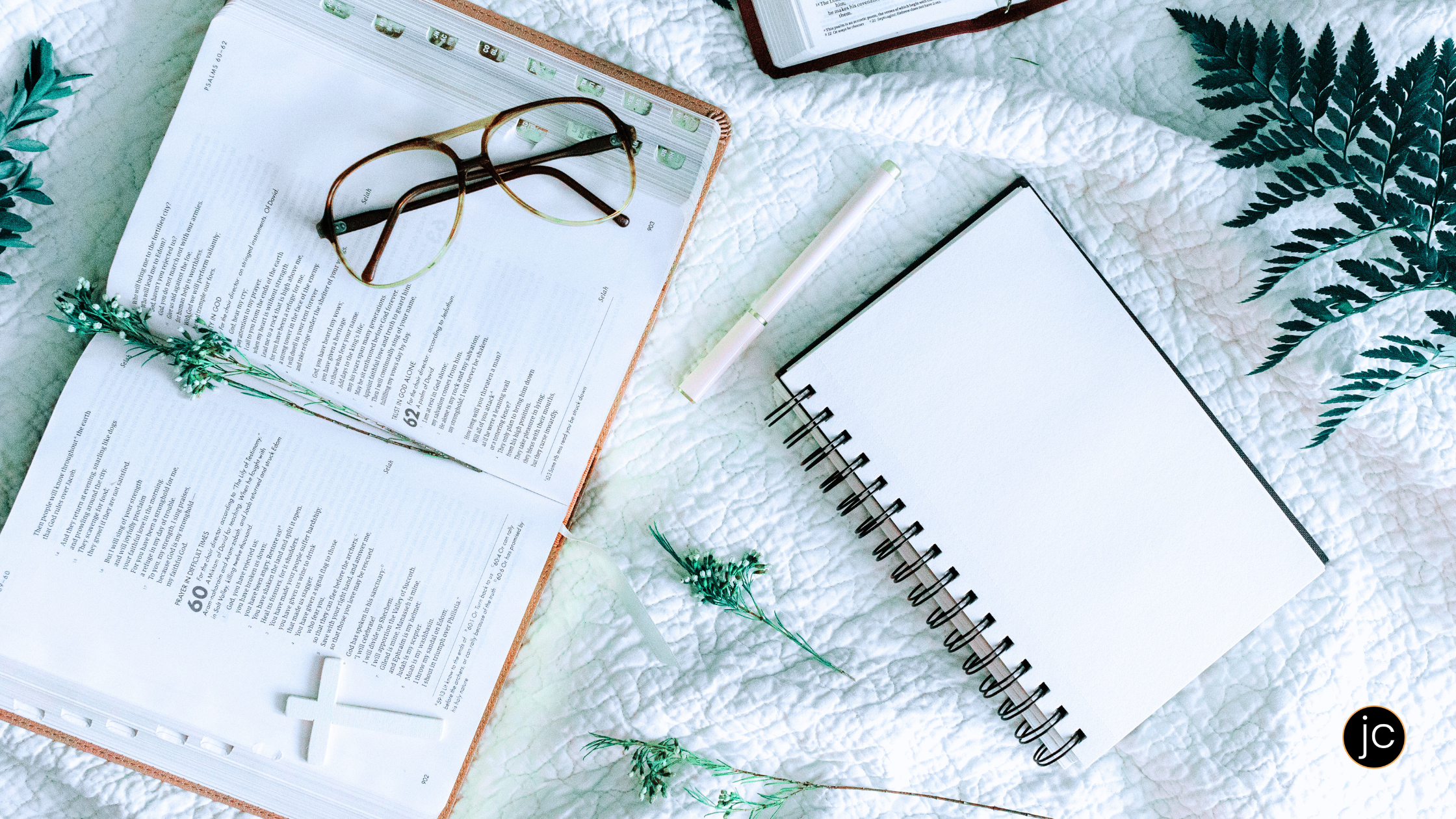 An open Bible with a pair of glasses laid on top, and an open notebook and a pen, all on a white quilt ready for someone to draw near to God through writing