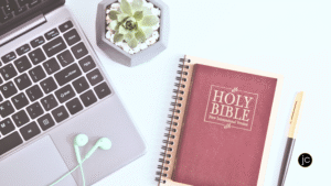 A laptop, ear buds, a succulent plant, a notebook, and a red Bible on a white desk