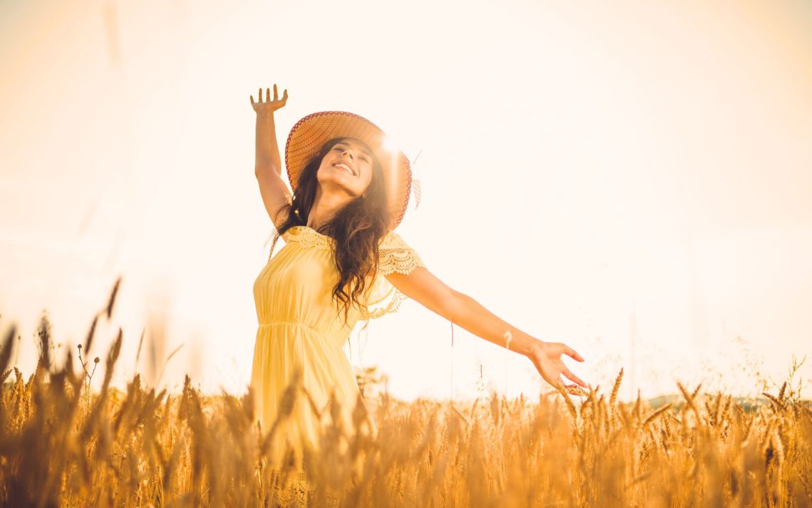 woman in straw hat and yellow sundress waving arms in a field of wheat at sunrise