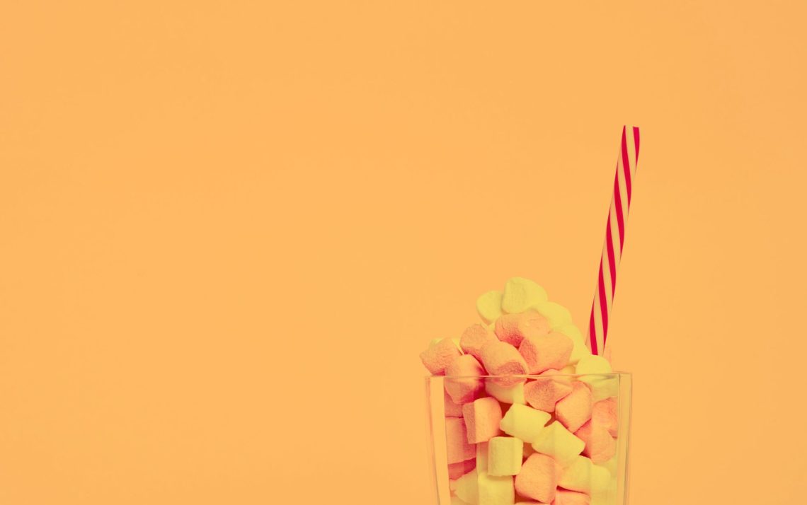 glass cup full of marshmallow candy with a striped red straw sticking out in front of a yellow background