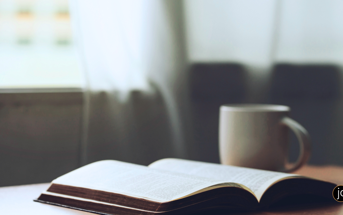 An open Bible and a steaming mug on a table in front of an open window in the morning light